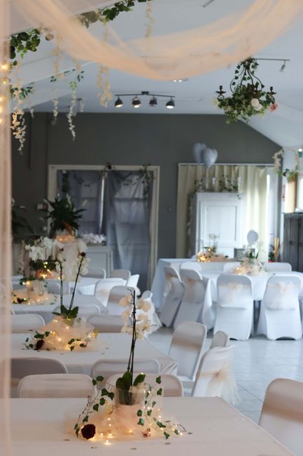 An elegantly decorated floral arrangement featuring white orchids and dark burgundy roses, placed in a transparent glass vase on a table during a wedding reception setup. The arrangement is surrounded