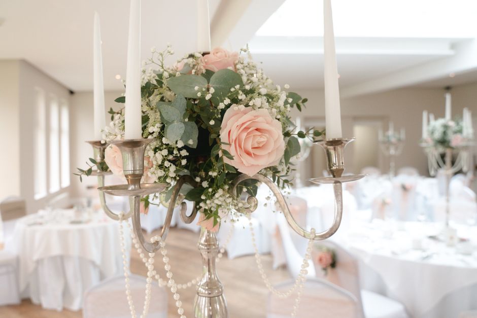 A floral centerpiece featuring soft pink roses, white baby's breath, and green eucalyptus leaves arranged in a silver candelabrum with three arms, adorned with hanging pearl strands. The bouquet is se