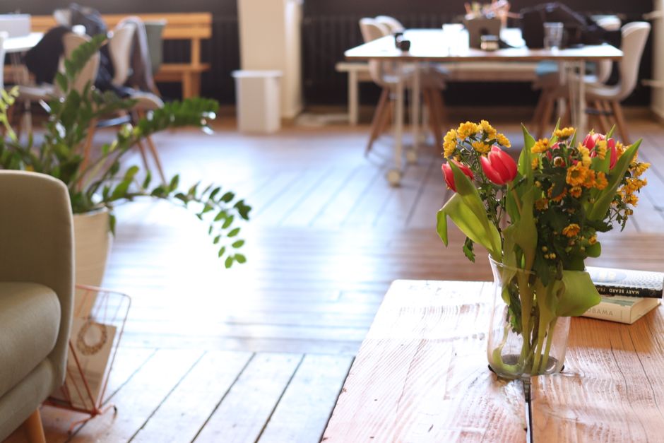 A clear glass vase filled with a vibrant floral arrangement featuring pink tulips, yellow daisies, and orange marigolds sits on a wooden table inside a well-lit, modern office space. The arrangement s