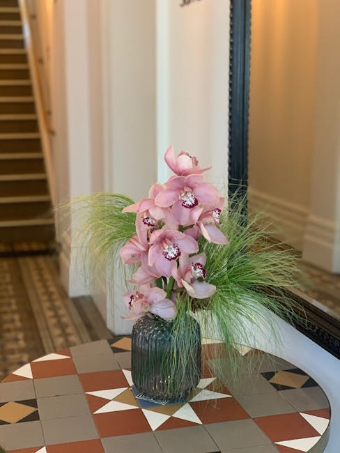 A tall, elegant floral arrangement featuring pale pink orchids with multiple blooms on each stem, displayed in a dark, ribbed glass vase. The orchids are complemented by wispy green grasses that fan o