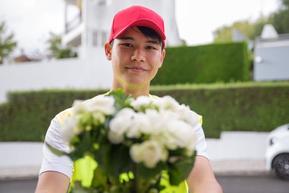 A delivery person with a friendly expression, wearing a red cap and a high-visibility vest, hands over a floral bouquet consisting of white roses, ranunculus, and greenery, wrapped in clear and white 