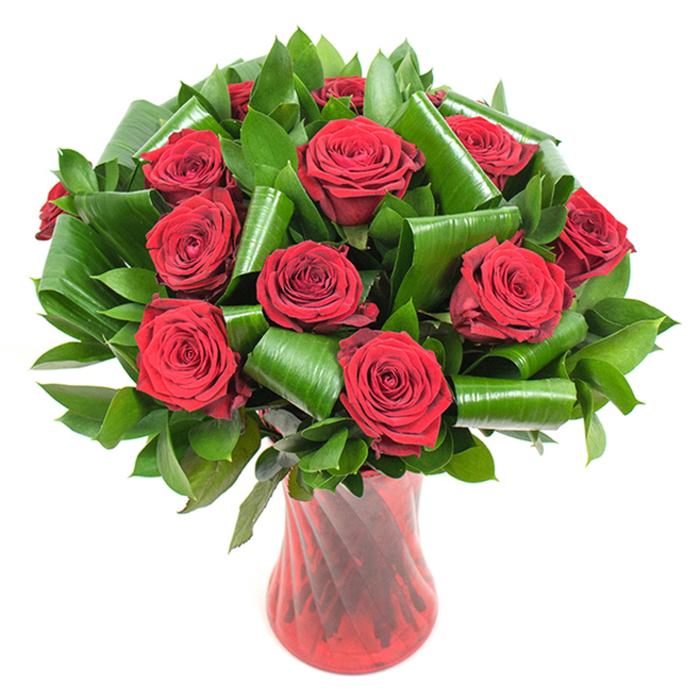 Here is a carefully composed, museum-like presentation of deep red roses - about a dozen or slightly more - where every flower reads as fully opened and luxuriously full, their velvety petals layered in tight spirals that speak of freshness and meticulous selection. The roses are interspersed with a generous quantity of dark, glossy foliage that provides both volume and visual rest; among these greens are broader, shining leaves that have been rolled and folded into sculptural cylinders, their edges coiling into architectural forms that cradle the blooms and introduce a contemporary silhouette. The stylistic contrast between the soft, tactile rose heads and the structured, almost origami-like foliage creates depth and rhythm across the bouquet. Supporting stems settle into a tall, slender glass vase moulded in a deep ruby tone - translucent yet richly coloured - with a subtle twisted or ribbed surface catching the light in vertical highlights that echo the stems' upward motion. The scene is lit with soft, even illumination against a pristine white backdrop, which isolates the composition and allows the deep crimson and emerald greens to saturate the eye without distraction. The overall expression is classic romance reinterpreted with modern lines, perfect for anniversary tables or elegant Valentine's gestures, and it carries local charm suited to Muswell Hill's neighbourhood gatherings near Muswell Hill Broadway or a private moment after a visit to Alexandra Park. The photograph conveys tactile cues - the plushness of petals, the cool gloss of leaves, and the smooth heft of glass - offering a sensory portrait of floral craft and refined, heartfelt intention.