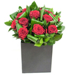 In this crisply modern composition, deep crimson roses form the heart of a compact, architectural bouquet-each bloom velvety and full, their petals unfurling in neat, generous spirals that catch the light with a soft, velvet sheen. Broad, glossy green leaves have been thoughtfully curled and interwoven around the roses, creating a rhythmic pattern of movement and contrast: the leaves' smooth, almost waxy surfaces reflect highlights while their edges loop to suggest motion. The flowers sit low and confident in a matte black square vase, a cube of quiet strength that grounds the arrangement and sets the vivid reds and greens against an urbane backdrop. Lighting here is clear and steady, as if photographed on a bright Muswell Hill morning near Alexandra Park, so every leaf and petal reads with crisp clarity. Bring the bouquet closer and you can almost sense the scent-clean, high notes of fresh foliage mingling with the warm, heady perfume of classic red rose-an olfactory echo of thoughtful intent. As a local florist would note, the craftsmanship is deliberate: foliage folded to sculpt, roses positioned to sing together, and negative space carefully respected so the design breathes. This arrangement suits contemporary urban interiors or a loved one's anniversary, offering restrained drama without excess. Delivered across Muswell Hill and nearby streets, it feels like a confident, affectionate gesture-both an objet d'art and a tender message, ready to transform a modern flat on Muswell Hill Broadway or a dinner table near Fortis Green Road into something quietly memorable.